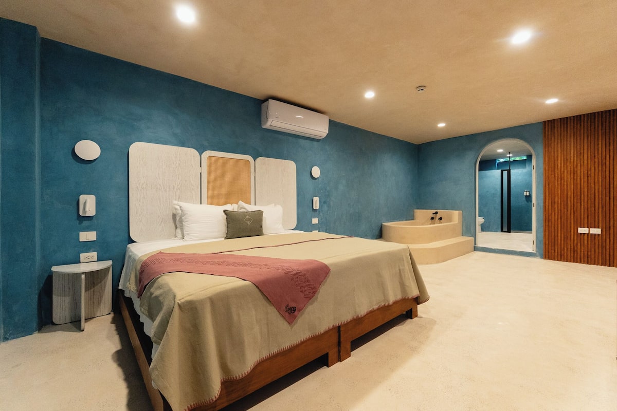 A bedroom with a large bed, a bedside table, a nightstand, an air conditioning unit, and a bathroom with a bathtub and shower.