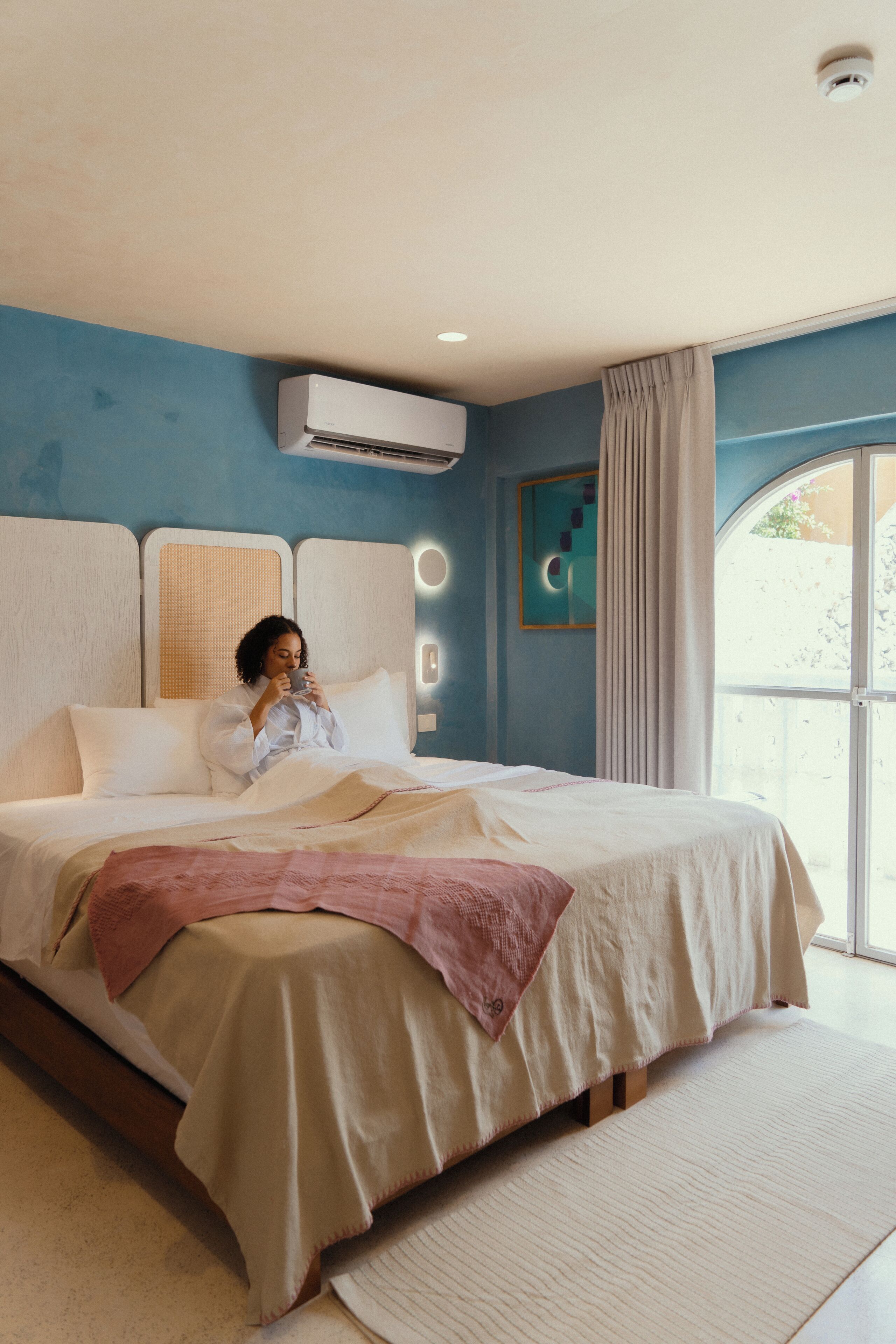 A bedroom with a large bed, an air conditioning unit, a window with curtains, and a wall-mounted light fixture.