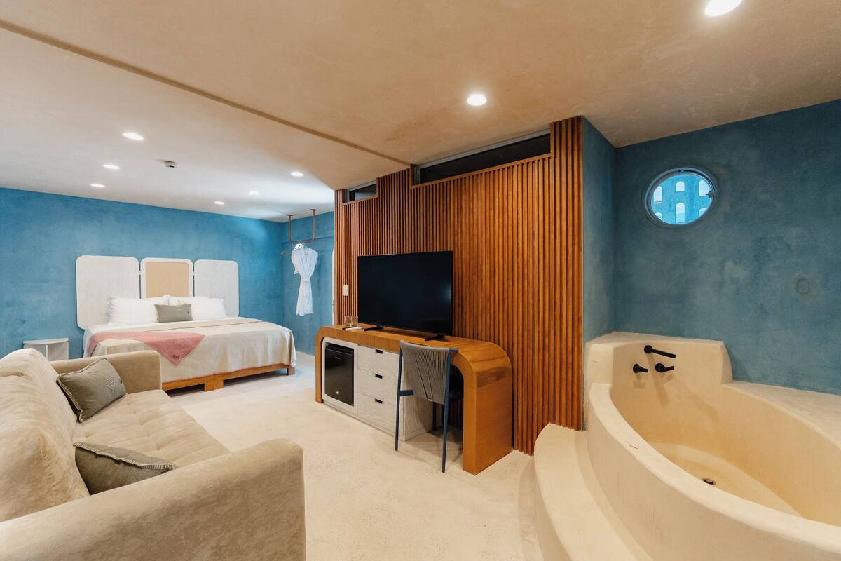 A modern bedroom with a large bed, a TV, a wooden entertainment unit, a bathtub, and a blue accent wall.