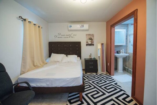 A bedroom with a bed, a nightstand, a wall-mounted air conditioner, and a bathroom visible through an open door.
