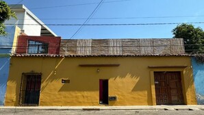 Un bâtiment jaune avec une porte rouge et deux fenêtres, une petite enseigne au-dessus de la porte et un toit en tôle ondulée.