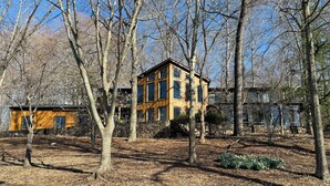A two-story wooden cabin with a stone foundation, surrounded by trees and a stone wall.