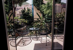 Un balcon avec des chaises et des tables en métal, une vue sur des immeubles et des arbres, et un ciel dégagé.