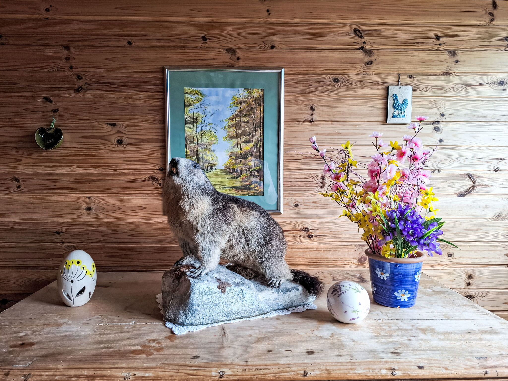 A wooden wall with a framed painting, a ceramic rooster, a vase with flowers, and a painted egg.