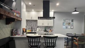 A modern kitchen with white cabinets, a black island, two bar stools, a dining area with a table and chairs, and a herringbone-patterned backsplash. - 2 Bedroom 2 Bath Condo (Tulsa)
