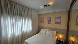 Una habitación de hotel con una cama, un banco, una mesita de noche con lámpara y tres cuadros en la pared.