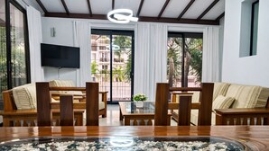 A modern hotel room with a wooden dining table, two sofas, a TV, and large windows with a view of buildings and greenery.