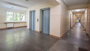 A long, narrow hallway with two blue doors, a radiator, and a window with a view of trees.