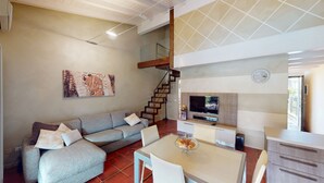 A modern living room with a grey sofa, a dining table with chairs, a staircase, a flat-screen TV mounted on a wooden panel, and a painting on the wall. - Maison Belvedere By Bookinggardalake (Manerba del Garda)