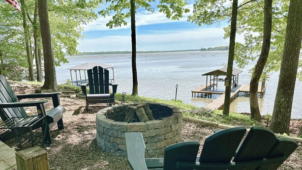 A lakeside deck with Adirondack chairs, a stone fire pit, and a wooden dock extending into the water. - Lake House Cabin 3BR/2BA Sleeps 6 Water Front With Private Boat Dock for Fishing (Cedar Bluff)