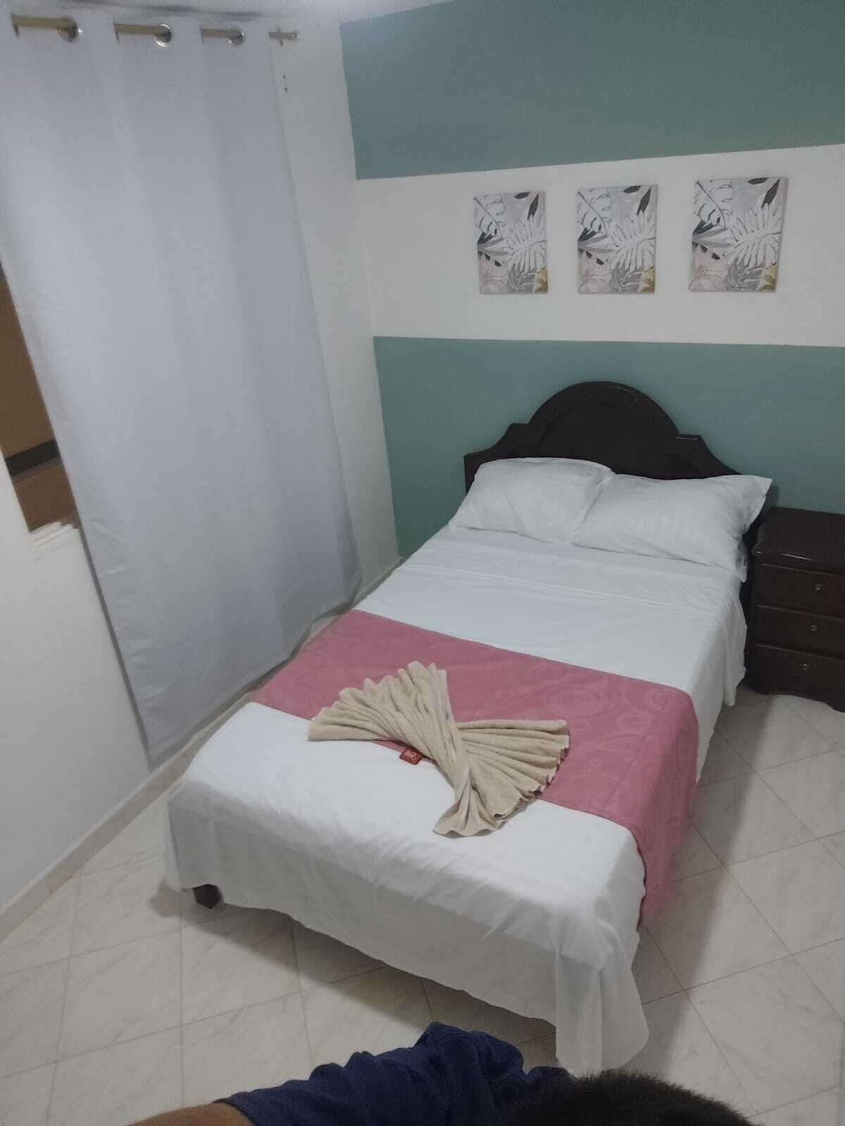 Quarto com cama, mesa de cabeceira, cortina e três quadros na parede.