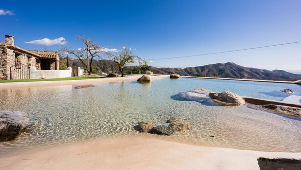 A clear pool with rocks, a stone building, and mountains in the background. - Cortijo Rural Fandango 3 - Pool and Mountain (Almuñecar)