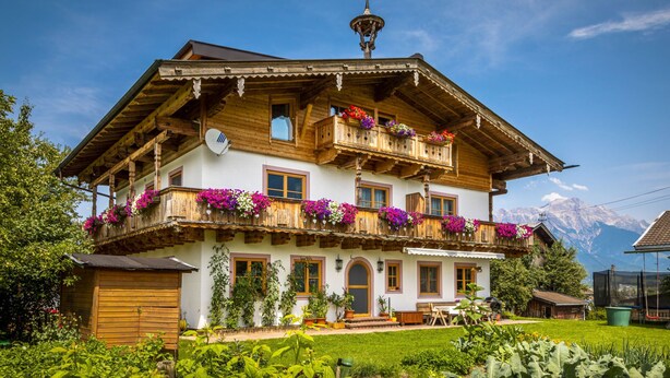 A two-story wooden house with balconies adorned with flowers, a garden in the foreground, and a mountainous backdrop.