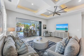 A modern living room with a large TV, comfortable seating, and a view of the outdoors. - New! Seashell Oasis - 3 Ensuites! 1 Mile to the Beach! - Heated Pool! (Naples)