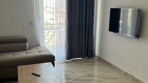 A living room with a sofa, a coffee table, a flat-screen TV mounted on the wall, and a large window with curtains.