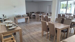 A spacious dining area with multiple tables and chairs, a wooden floor, and large windows allowing natural light.