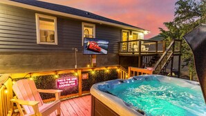A hot tub on a wooden deck with a TV screen displaying a sports car, a neon sign, and a staircase leading to a second level.