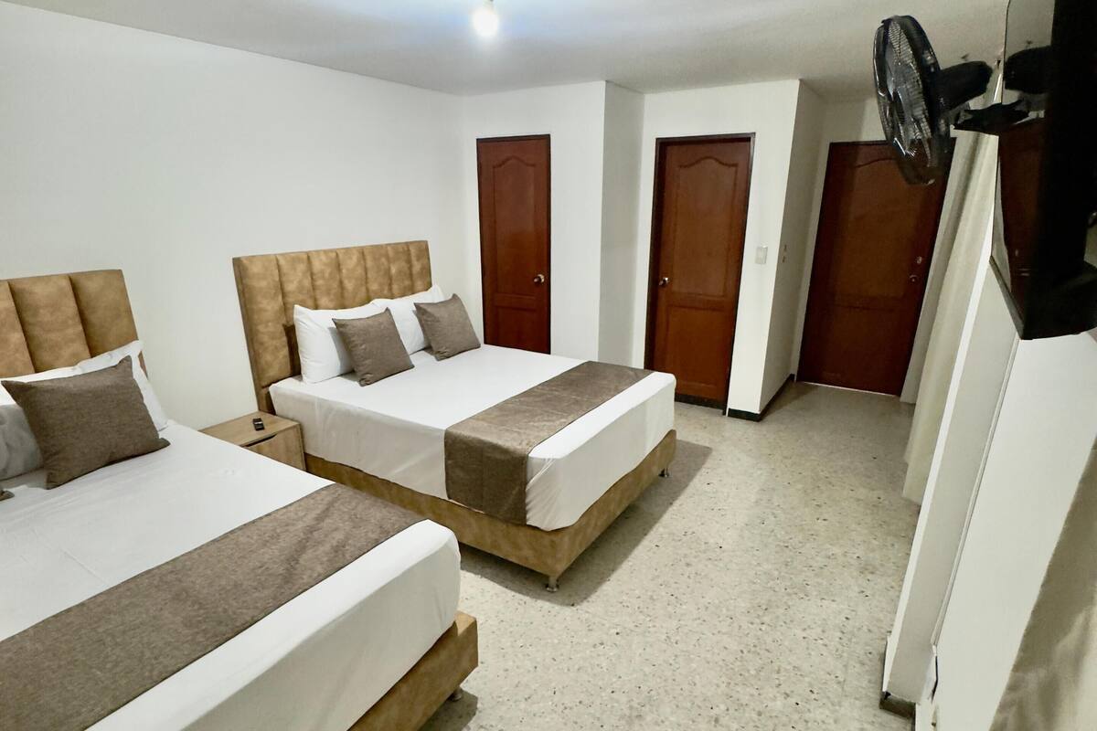 Quarto de hotel com duas camas, um ventilador de teto e uma televisão fixada na parede.