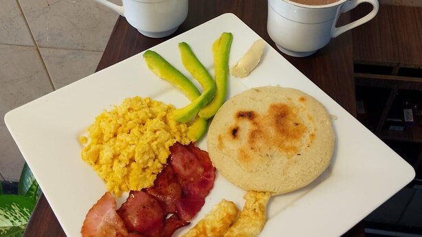 A breakfast plate with scrambled eggs, bacon, avocado slices, and a arepa, accompanied by two cups of coffee and a bottle of honey. - Hotel Pereira Central (Pereira)