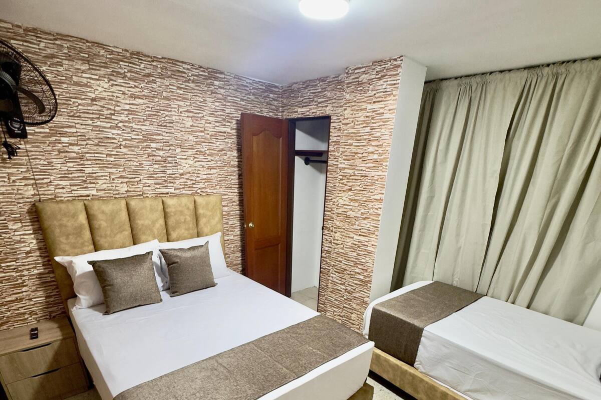 Quarto de hotel com uma cama grande, uma cama menor, uma mesa de cabeceira, um guarda-roupa e um ventilador de teto.