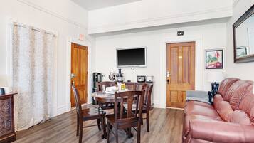 A dining area with a wooden table and chairs, a television mounted on the wall, and a couch with a cushion.