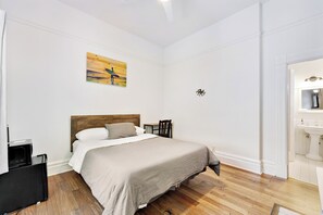 Un dormitorio con cama, una mesita, una silla, un cuadro y un baño con lavabo y espejo.