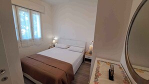 A bedroom with a bed, two bedside tables with lamps, a window with blinds, and a mirror on the wall.