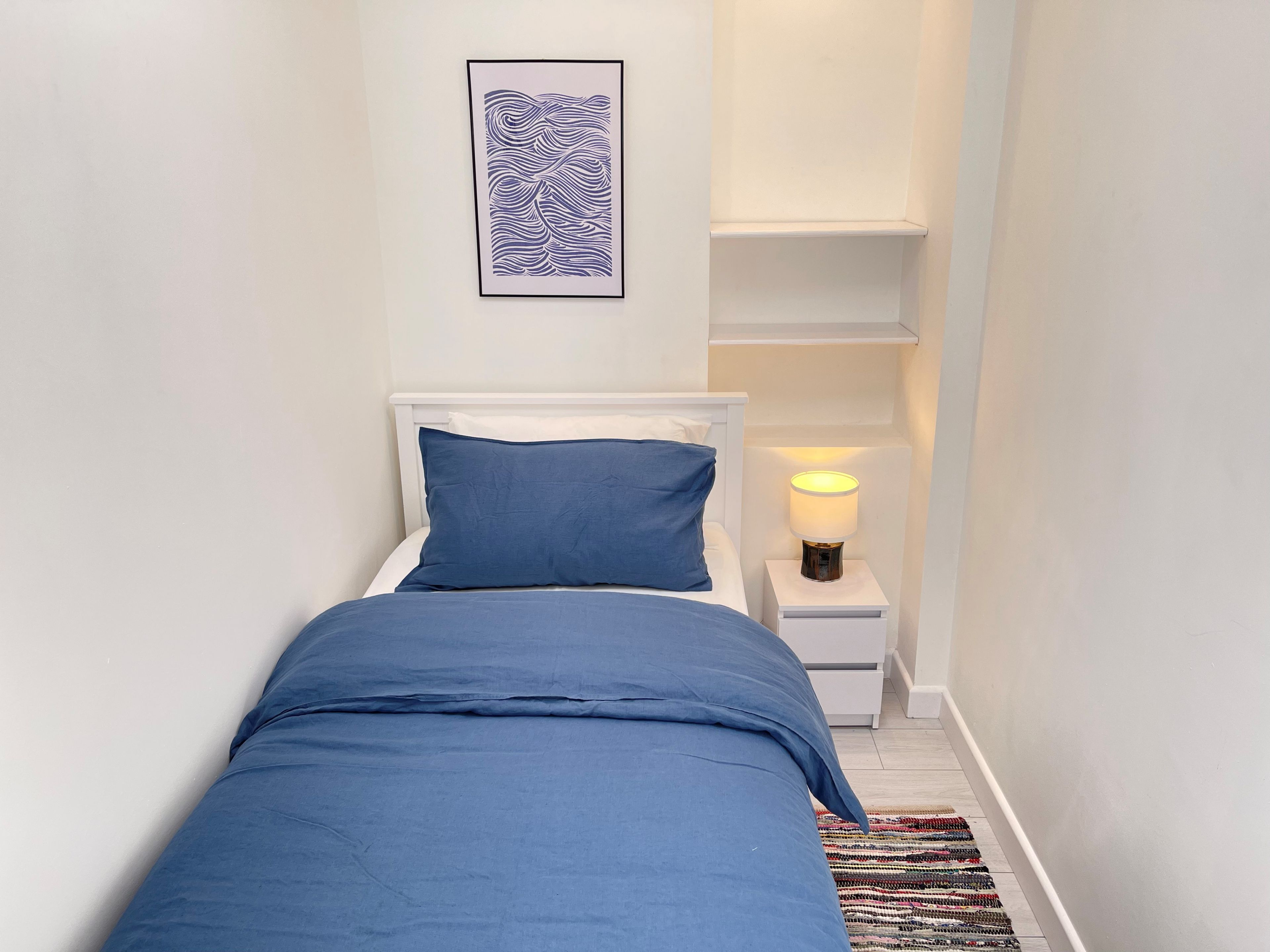 Ein schmaler Flur mit einem Bett, ausgestattet mit einer blauen Tagesdecke und Kissen, einer Nachttischlampe und einem gerahmten Kunstwerk an der Wand.