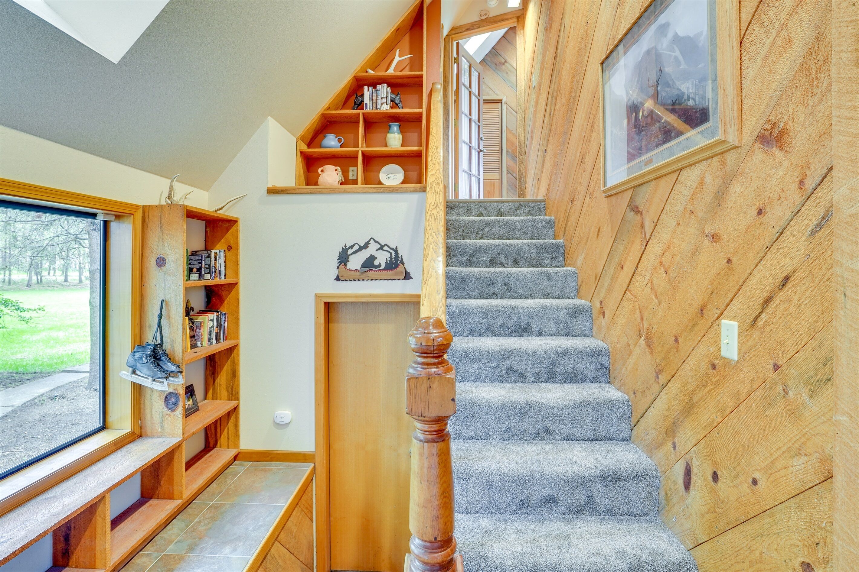 Una escalera de madera con rellano alfombrado, estantes de madera con libros y objetos decorativos, un cuadro enmarcado en la pared y vistas a través de una ventana.