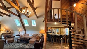Een loft-achtige woonruimte met een hoge nok, zichtbare houten balken, een trap en een keuken op de achtergrond.