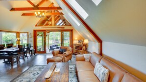 Een ruime loft met hoge plafonds, zichtbare houten balken, een eethoek met tafel en stoelen, een leren bank en een groot raam met uitzicht op het omliggende landschap.