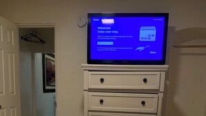 Une chambre d’hôtel avec un lit, une télécommande, un téléviseur affichant l’écran d’accueil de Roku, et un couloir avec une porte.