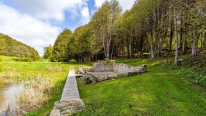 A stone pathway leading through a lush green landscape with a small body of water on the left and a dense forest on the right. - Exclusive 100-Acre Modern Estate Designed Home (Alton)