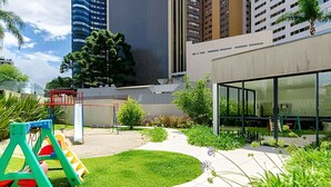 A modern building with a glass facade, a playground with a slide, and a well-maintained garden area. - 1 quarto, cheio de estilo, condo club Sky com vaga (Curitiba)