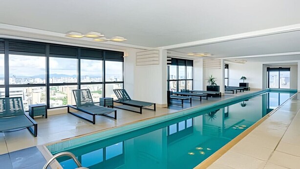 A modern indoor swimming pool with clear glass walls, multiple lounge chairs, and a city view. - 1 quarto, cheio de estilo, condo club Sky com vaga (Curitiba)