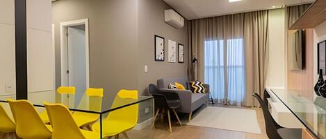 A modern living room with a glass dining table, yellow chairs, a grey sofa with black and yellow cushions, and a large window with curtains.
