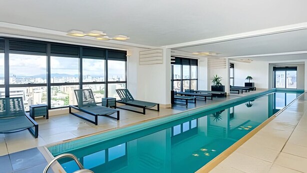 A modern indoor swimming pool with clear views of the city through large windows. - Oásis Urbano com Vistas Deslumbrantes no Sky CWB (Curitiba)