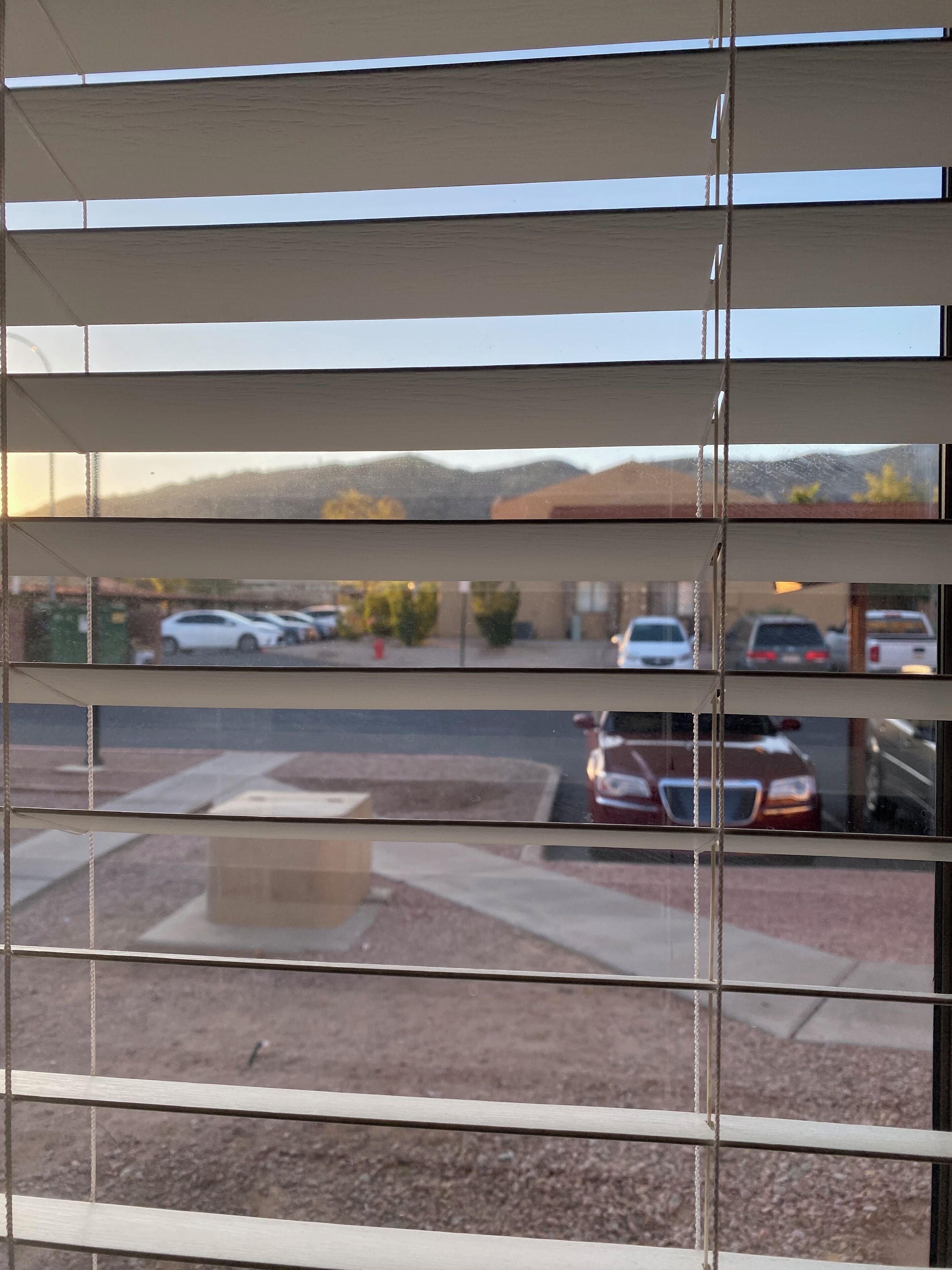 A view through a window with horizontal blinds, revealing a parking lot with several cars and a building in the background.
