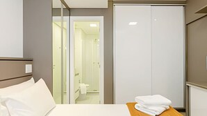 Habitación de hotel con cama, una mesita de madera con toallas, un armario y un baño visible a través de una puerta de cristal.