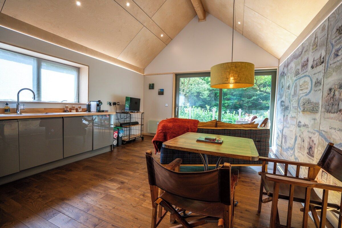 A modern kitchen with a dining area, wooden flooring, and a large window providing a view of greenery.