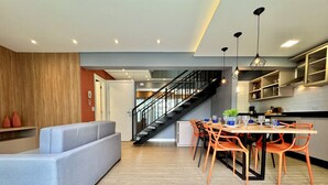 A modern living room with a grey sofa, a dining area with orange chairs, a staircase, and a kitchen with wooden cabinets. - Duplex Incrível, 2 Quartos, varanda, 2 vagas Batel (Curitiba)