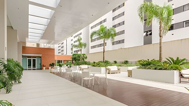 A spacious indoor area with a wooden floor, white walls, and a high ceiling with skylights. There are several white chairs and tables arranged for outdoor dining. - Duplex Incrível, 2 Quartos, varanda, 2 vagas Batel (Curitiba)