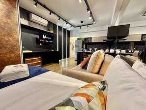 A modern hotel room with a bed, a sofa, a TV, a kitchen area, and an air conditioning unit.