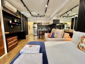 A modern hotel room with a flat-screen TV, a sofa, a bed, a desk, and a kitchenette. - Studio reformado no Batel para 4, piscina aquecida e Shopping Curitiba em frente (Curitiba)