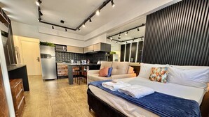 A modern bedroom with a large bed, a kitchenette in the background, and a wooden floor.