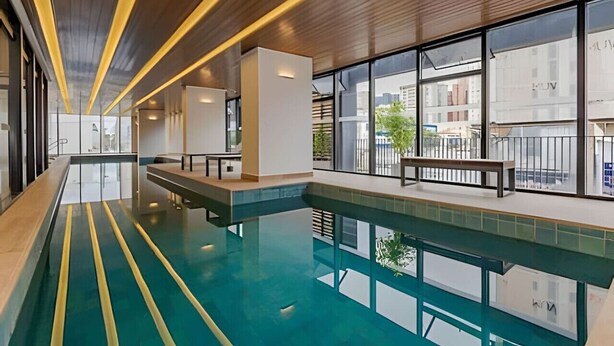 A modern indoor swimming pool with clear glass walls, wooden ceiling, and a bench along the poolside.