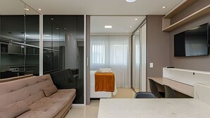 A modern hotel room with a sofa, a bed with a white cover, a desk, and a TV mounted on the wall.