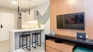 A modern kitchen with a white countertop, black bar stools, a wooden cabinet, and a flat-screen TV mounted on the wall. - Apto charmoso de 1 quarto mais sala ed. Elysia (Curitiba)
