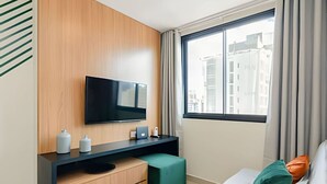 A modern hotel room with a large window, a flat-screen TV mounted on a wooden panel, a small desk with a laptop, and a green ottoman. - Apto charmoso de 1 quarto mais sala ed. Elysia (Curitiba)