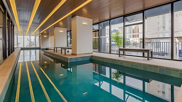 A modern indoor swimming pool with clear glass walls, wooden ceiling, and a bench along the edge. - Apto charmoso de 1 quarto mais sala ed. Elysia (Curitiba)